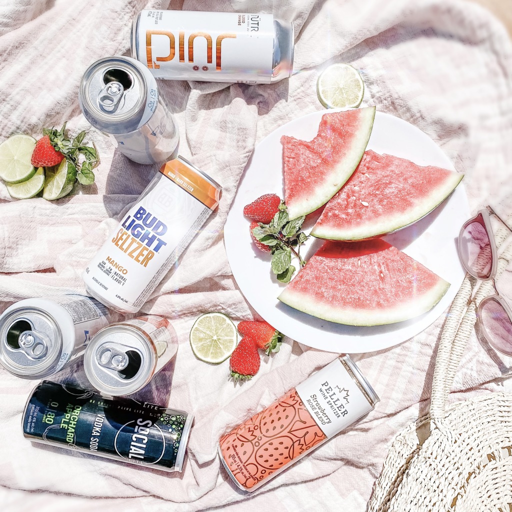 Low-Calories Summer Alcoholic Coolers laid on a picnic blanket with fresh fruit. Low-Calorie 2021 Summer Alcoholic Drink Review: The best low-sugar alcoholic coolers available in stores now at the LCBO. 